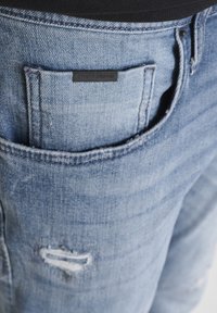 Jeans de mezclilla celeste con textura desgastada, que cuentan con una etiqueta en el bolsillo trasero que dice "ANTHONY MORATO" y un pequeño desgarro como detalle.