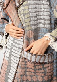 Tkani obavijen s mješavinom sive, plave i pastelnih uzoraka, s izfrkanim rubovima i dva prednja džepova sa prugastim detaljima.