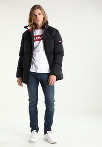 Schwarze Pufferjacke mit hohem Kragen, roten Akzenten und Reißverschluss, getragen über einem weißen Levi's Logo-T-Shirt und blauen Jeans. Weiße Sneaker.