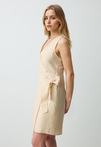 Beige wikkeljurk van lichtgewicht stof, met een diepe V-hals, mouwloze uitvoering en een strikaccent aan de linkerkant.