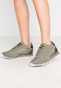 Baskets vert olive avec un mélange de matériaux lisses et texturés, dotées de lacets marron clair, d'une semelle blanche et de touches noires discrètes.