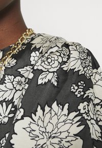 Czarna tkanina z białym kwiatowym wzorem, o teksturowej powierzchni. Widać złoty łańcuszek w okolicy dekoltu.