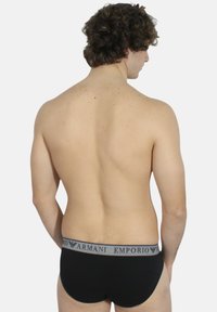 Slip in cotone nero con un elastico grigio, visti da dietro. Il tessuto appare liscio, con una vestibilità aderente e un design classico.