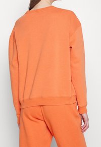 Felpa arancione con vestibilità rilassata, spalle scese, polsini e orlo a coste. Il tessuto sembra morbido e strutturato. Indossata con pantaloni abbinati.