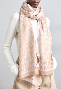 Beige scarf med upprepat "MK" mönster, tillverkad av mjukt material; har fransiga ändar och en lätt, texturerad design.