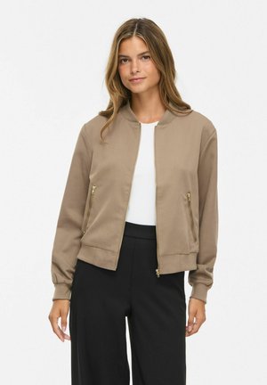 Femme portant une veste zippée marron clair sur une chemise blanche et un pantalon noir, debout devant un fond clair uni.
