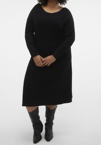 Robe noire tricotée à manches longues, coupe ample, avec un décolleté plongeant, associée à des bottes noires hauteur genou à bout pointu.