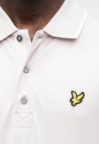 Lichtroze poloshirt met een gestructureerde stof, voorzien van een zwart geborduurd logo van een vogel met gele accenten en zwarte knopen.