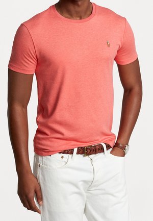 Homme portant un t-shirt corail à manches courtes et col rond, un pantalon blanc avec une ceinture tressée marron, une montre au poignet et la main dans la poche.