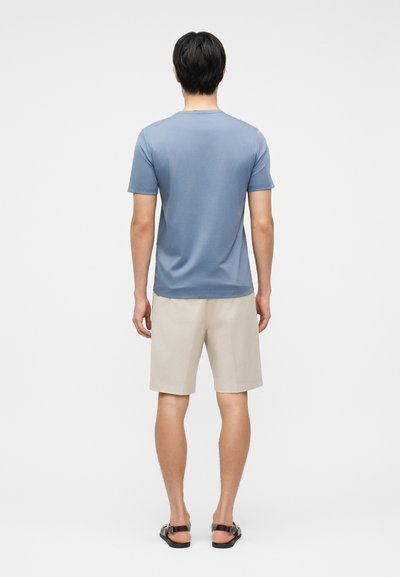 Άνδρας που στέκεται με την πλάτη προς τα εμπρός, φορώντας μπλε κοντομάνικο πουκάμισο, μπεζ σορτς και μαύρα σανδάλια σε απλό λευκό φόντο.