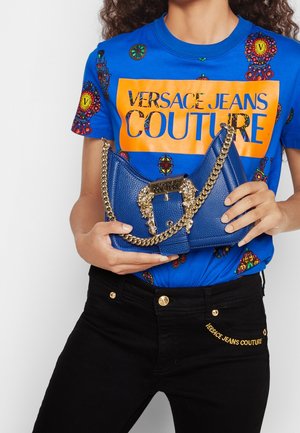 Chemise bleue Versace Jeans Couture avec des motifs colorés et un logo orange, tenant un sac à main bleu texturé avec une chaîne dorée et une boucle décorative.