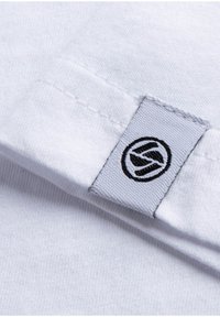 T-shirt bianco in cotone con colletto ripiegato. Presenta un'etichetta in tessuto grigia con un logo grafico nero ricamato su di essa. Texture liscia.