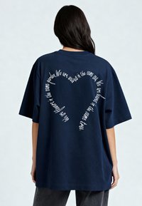 Tamsiai mėlynas per didelis medvilninis marškinėliai su balta tekstu širdies forma nugaroje, apvaliu iškirpimu ir trumpomis rankovėmis.