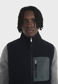 Gilet en polaire noir avec un col haut et des manches longues grises. Il dispose d'une poche zippée verte sur la poitrine gauche. Texture lisse et fermeture éclair à l'avant.