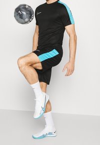 Schwarzes Sportshirt mit blauen Akzenten, passende schwarze Shorts und weiße Schuhe mit blauen Details; präsentiert einen schwarzen Fußball.