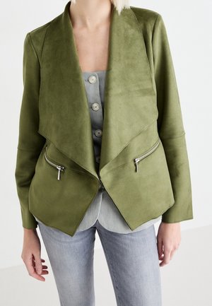 Mujer con chaqueta de ante verde oliva abierta con solapas anchas, bolsillos con cremallera y camisa gris claro abotonada debajo, combinada con vaqueros azul claro.