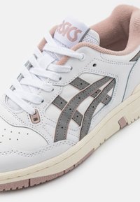 Zapatilla de cuero blanca con acentos en gris y rosa claro. Presenta una caja de dedos perforada, cordones y suela de goma texturizada. Logo de la marca en la lengua.