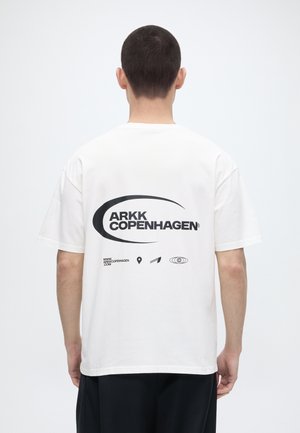 Bărbat cu păr scurt și închis la culoare, purtând un tricou alb ARKK Copenhagen cu logo negru și simboluri pe spate, văzut din spate.