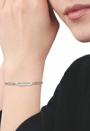 Pols met zilveren armband met ovale en rechthoekige schakels, waarvan één bezet met kleine heldere stenen, gedragen door een persoon met een zwarte mouw.