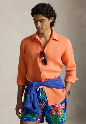 Mann trägt hochgekrempeltes orangefarbenes Hemd mit Knöpfen, blaue florale Badeshorts, einen blauen Pullover um die Taille gebunden und eine Sonnenbrille, die am Hemd hängt.