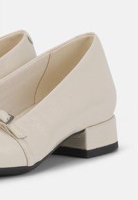 Beige fusk läderskor med låg klack, med en dekorativ rem i silver och en strukturerad yta.
