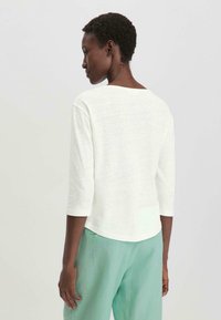 Weißes, leichtes Dreiviertelarmoberteil mit weitem Ausschnitt. Weiche Textur und lockere Passform, kombiniert mit hellgrünen Hosen.