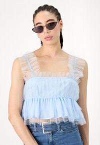 Világoskék, réteges crop top áttetsző tüllből, fodros pántokkal és testhezálló felsőrésszel, magas derekú farmernadrággal párosítva.