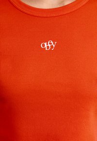 Ribbede rød T-skjorte med rund hals og liten hvit "obey"-logo på brystet. Materialet ser mykt ut med en teksturert overflate.