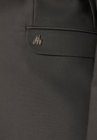 Tissu foncé avec une petite épingle monogramme en métal argenté présentant des lettres stylisées sur un rabat de poche d'un vêtement.