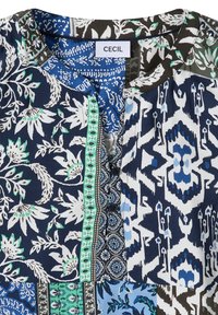 Blouse à col avec des motifs patchwork floraux, géométriques et abstraits en tons bleu, vert, blanc et noir, avec des boutons à l'avant.