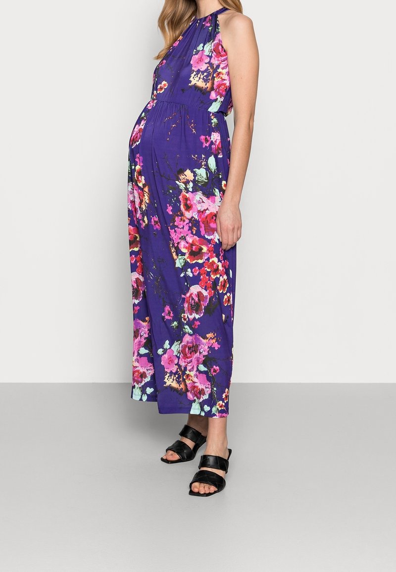 Femme enceinte portant une robe maxi fleurie violette sans manches et des sandales ouvertes noires, debout devant un fond blanc uni.