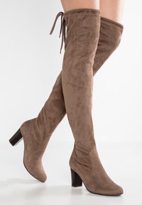 Botas marrones por encima de la rodilla en suave material de ante, con tacón grueso y detalle de lazo en la parte superior. Textura lisa y diseño ajustado.