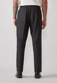 Pantalones negros ajustados con cintura elástica, que cuentan con bolsillos traseros y un tejido texturizado. Llevados con zapatos negros, vistos desde la parte de atrás.