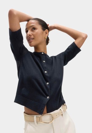 Frau richtet ihre Haare, trägt eine schwarze, kurzärmelige Strickjacke mit Knöpfen, beige hochtaillierte Hosen und einen passenden Gürtel mit goldener Schnalle.