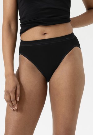 Frau trägt schwarze hochgeschnittene Slips und ein schwarzes, kurzes Oberteil, steht vor einem einfarbigen hellen Hintergrund.