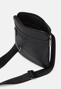 Černá kožená kabelka přes rameno s texturovaným povrchem, horním zapínáním na zip a nastavitelným popruhem. Má vnější kapsu na zip.