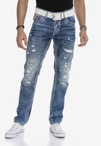 Blaue Jeans aus Denim mit distressed Details, weißem perforierten Gürtel und kontrastierenden Nähten. Kombiniert mit weißen Sneakern. Lässiges Passform-Design.