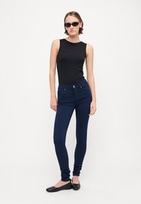 Svart ermeløs topp, mørkeblå tettsittende jeans, og svarte leiligheter. Modell som har på seg smale solbriller, står mot en enkel hvit bakgrunn.