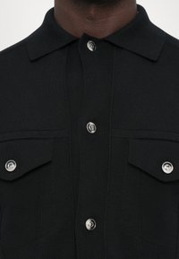 Černá, žebrovaná polokošile s límcem, obsahující dvě přední kapsy a stříbrné knoflíkové detaily. Strukturovaná tkanina s čistým provedením.