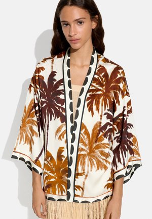 Kobieta nosząca kremowe kimono z brązowymi i pomarańczowymi nadrukami palm oraz czarną lamówką w groszki, do tego beżowa bluzka i spódnica z frędzlami.