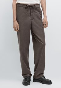 Pantalons marron sur mesure avec un tissu texturé, taille élastique à cordon, poches latérales et coupe droite décontractée. Portés avec des chaussures noires.