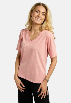 Blonde Frau lächelt, trägt ein hellrosa V-Ausschnitt Shirt mit kurzen Ärmeln und schwarze Hose, steht vor einem schlichten weißen Hintergrund.