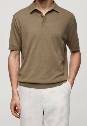 Mand iført en brun poloshirt med korte ærmer, stukket ned i lyse bukser, med en hånd i lommen mod ensfarvet baggrund.