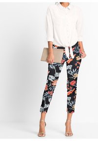 Chemise blanche à boutons nouée à la taille, associée à un pantalon noir à fleurs avec des motifs rouges, orange et bleus, et une pochette beige. Talons argentés.