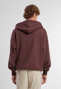 Tmavo hnedá oversize mikina vyrobená z mäkkého bavlneného materiálu, vybavená kapucňou na stiahnutie a ribbed manžetami, s hladkou, jednoduchou textúrou.