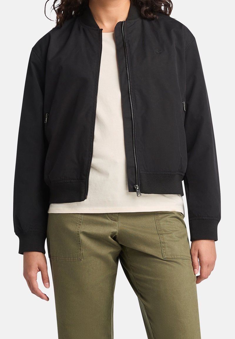 Personne portant une veste bomber noire zippée, une chemise beige et un pantalon vert olive avec des poches plaquées à l'avant, debout devant un fond blanc.
