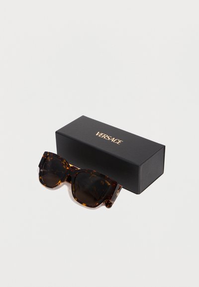 Lunettes de soleil surdimensionnées carrées écaille de tortue avec des verres foncés posées à côté d'un étui à lunettes noir marqué Versace sur un fond blanc.