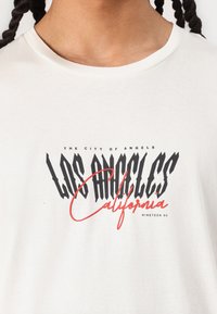 Off-white bomulds t-shirt med fed sort og rød tekst: "LOS ANGELES California," med et slidt grafisk design og rund hals.