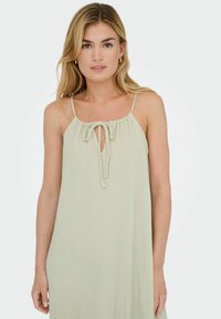 Donna con capelli biondi che indossa un vestito senza maniche di colore verde chiaro con spalline sottili e un collo con apertura a forma di chiave, in piedi su uno sfondo bianco uniforme.