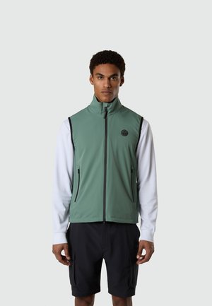 Man wearing sleeveless green zip-up vest over white long-sleeve shirt and black cargo shorts, standing against plain light background.